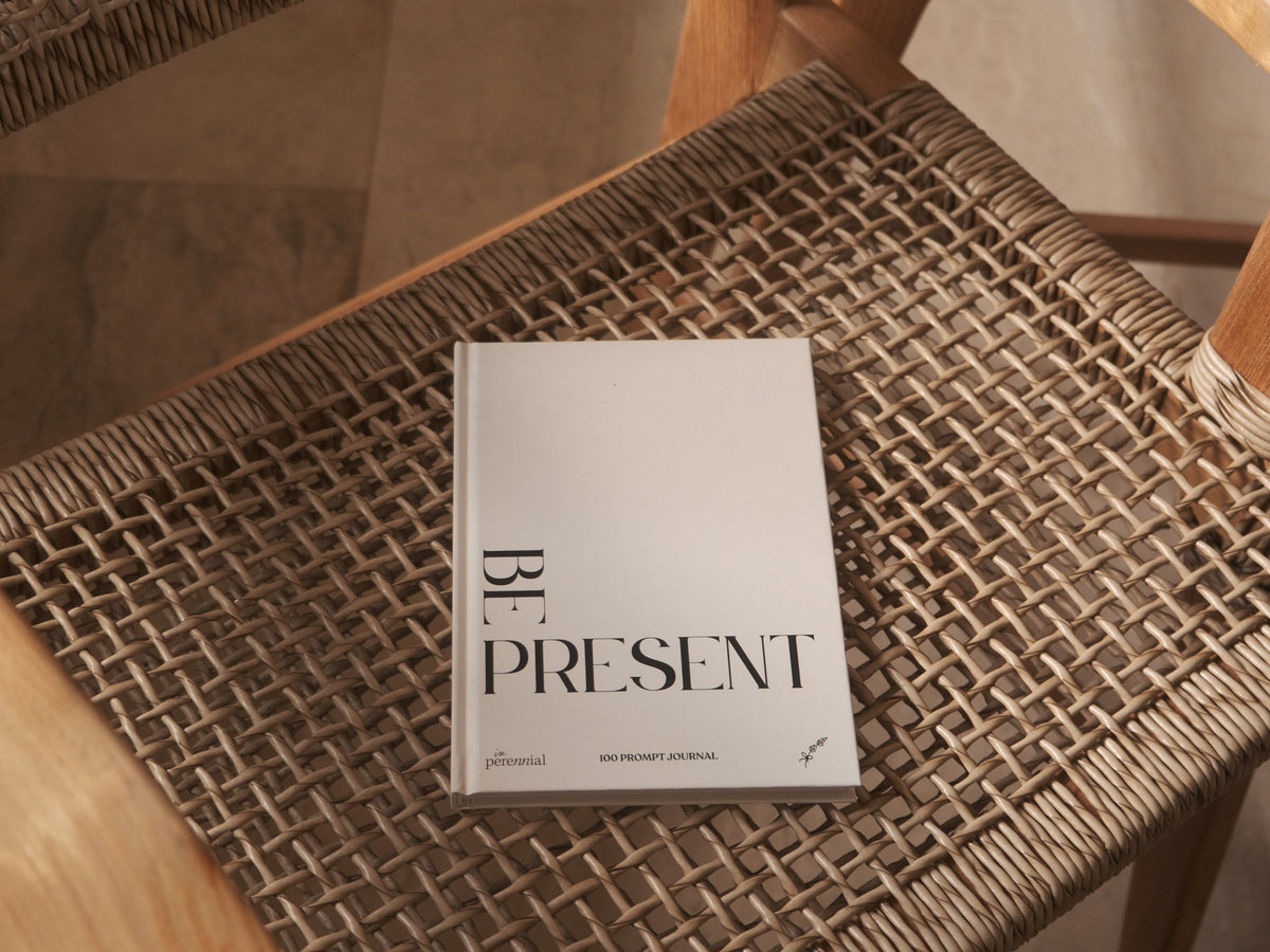 Be Present guided journal by in Perennial on a woven chair seat, with its beige front cover and black lettering visible.